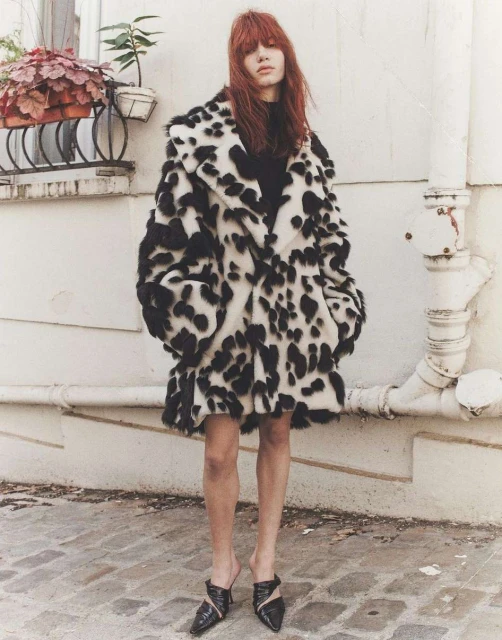 A fashionable adult female stands outdoors wearing a striking black-and-white spotted faux fur coat, black top, short skirt, and stylish mules.