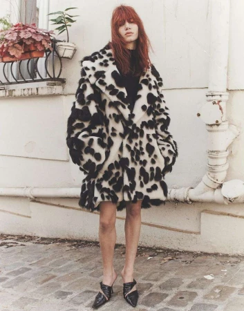 A fashionable adult female stands outdoors wearing a striking black-and-white spotted faux fur coat, black top, short skirt, and stylish mules.