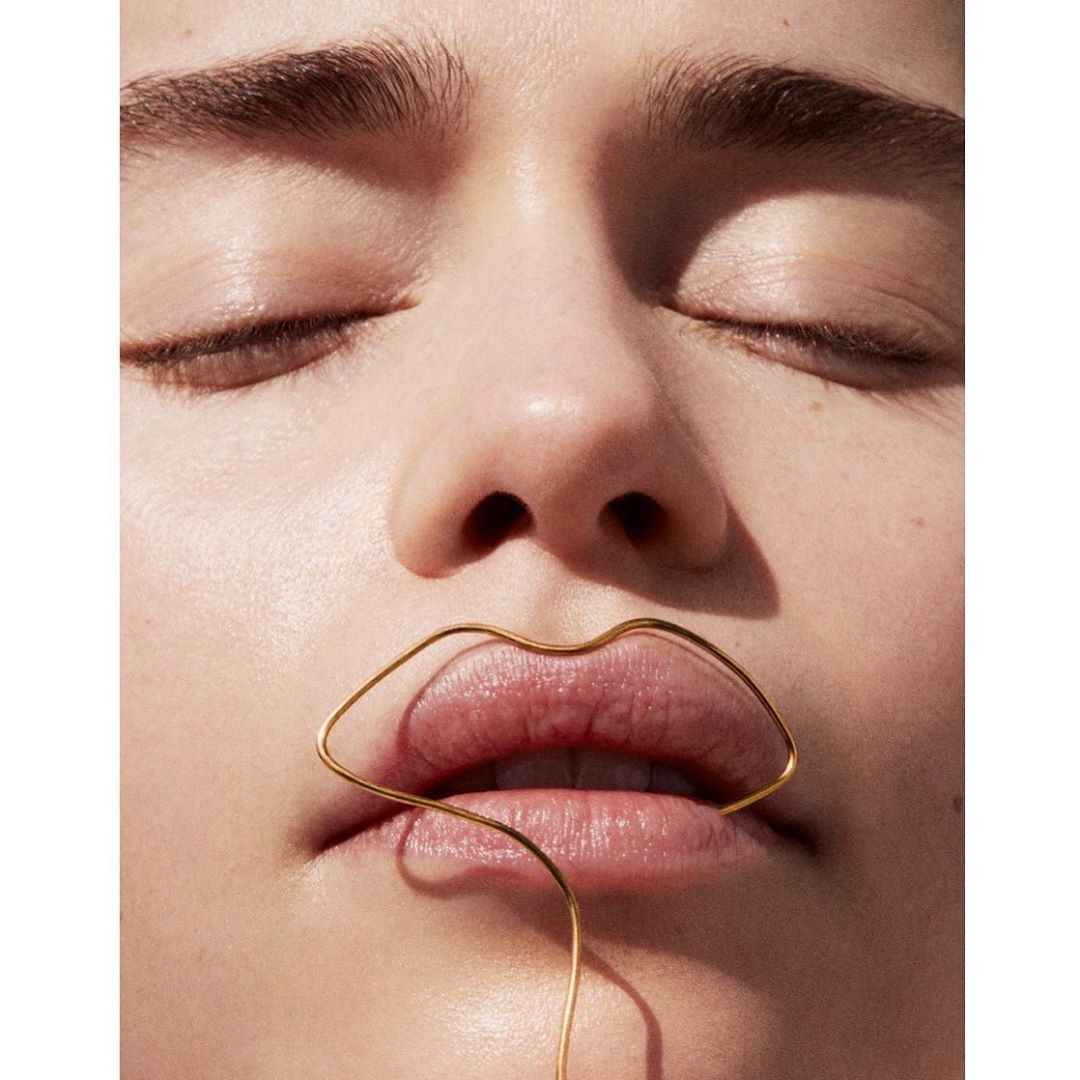Close-up of an adult woman's face with closed eyes and a gold wire sculpture of lips placed over her actual lips in a beauty shot.