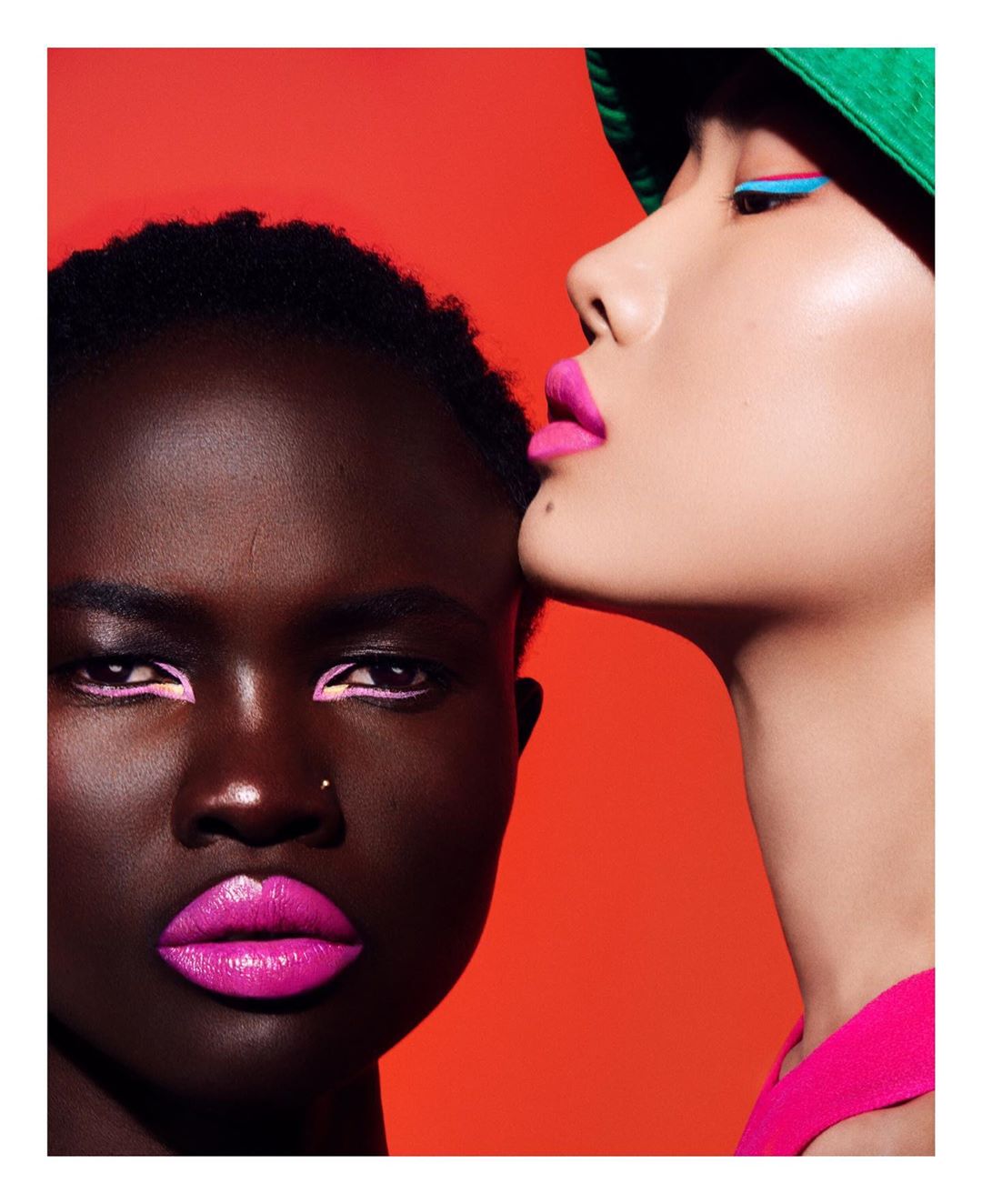 Close-up of two diverse women showcasing beauty makeup: vibrant pink lips, colorful eyeliner, and striking color contrast against an orange background.