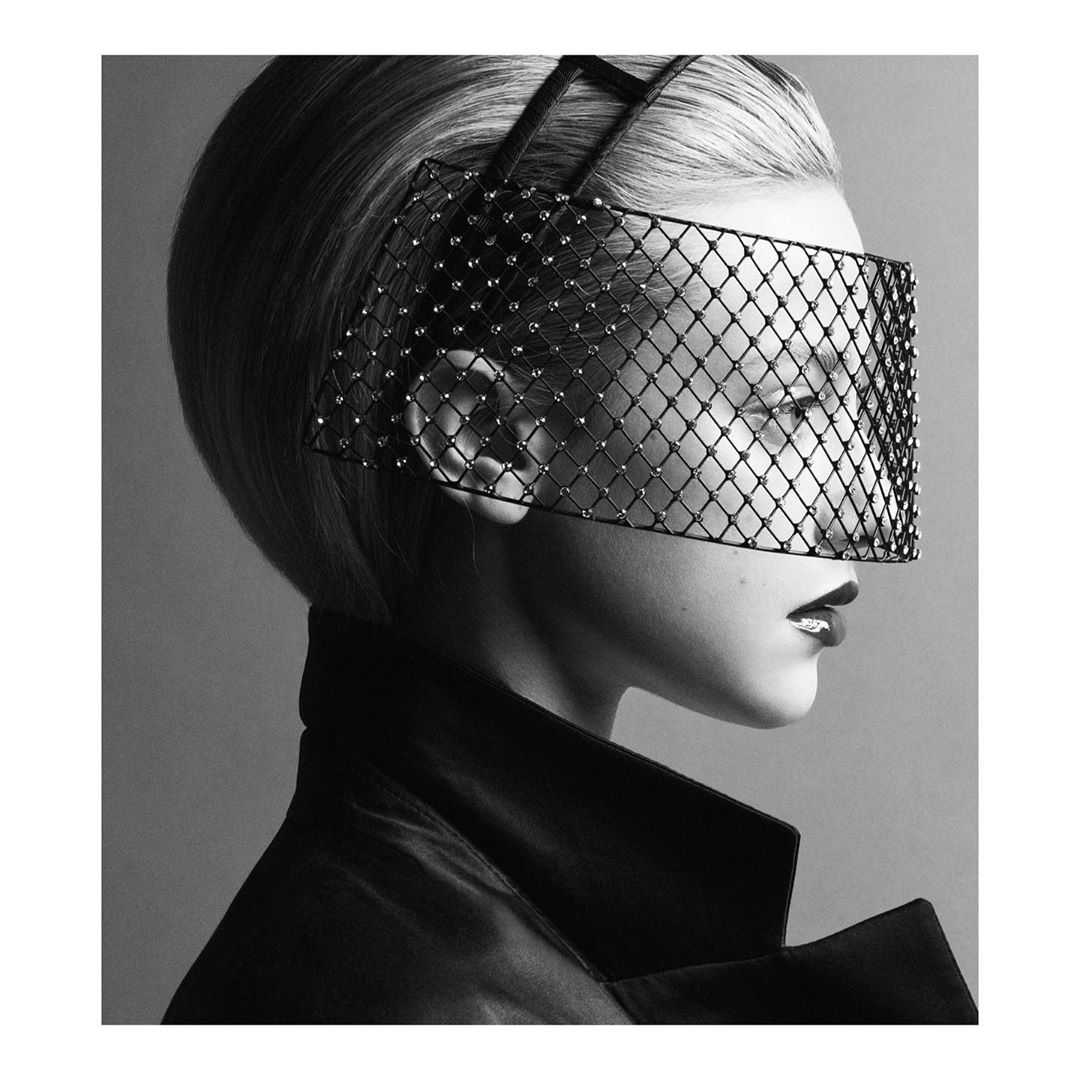 Monochrome portrait of a woman with short blonde hair, wearing a mesh-covered headpiece and dark coat, showcasing a high-fashion look.