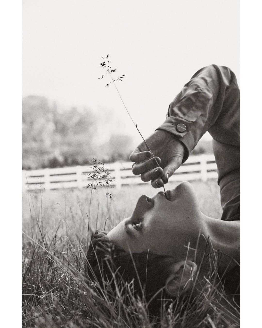 Person lying in a grassy field, holding a stem to their mouth, creating a serene, monochromatic outdoor portrait in natural lighting.