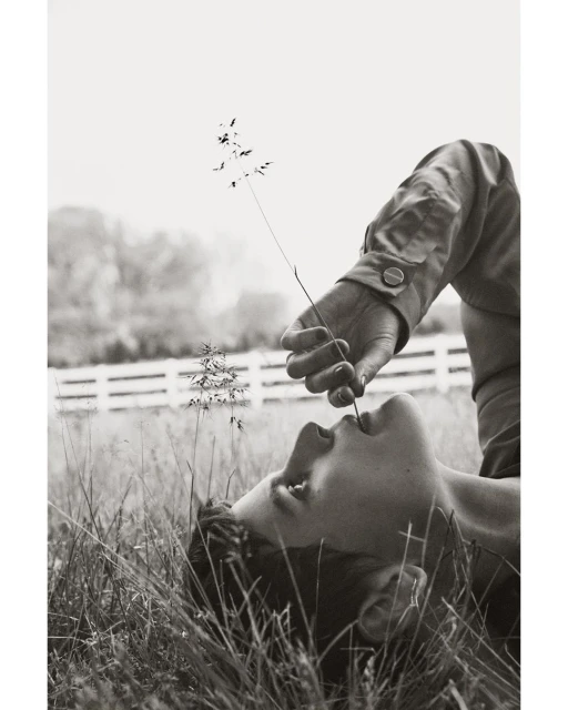 Person lying in a grassy field, holding a stem to their mouth, creating a serene, monochromatic outdoor portrait in natural lighting.