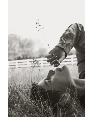 Person lying in a grassy field, holding a stem to their mouth, creating a serene, monochromatic outdoor portrait in natural lighting.