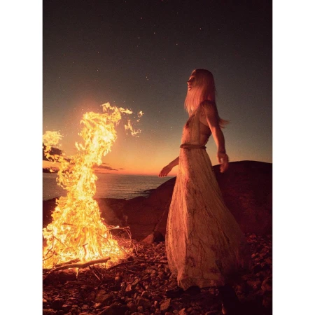 Blond woman in dress stands by large bonfire on rocky coast at dusk with orange-lit horizon, and starry sky above.
