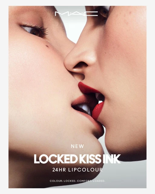 Close-up of two adult women kissing, featuring red lipstick and 'MAC' logo. Beauty advertisement with red lips and fair skin tones.