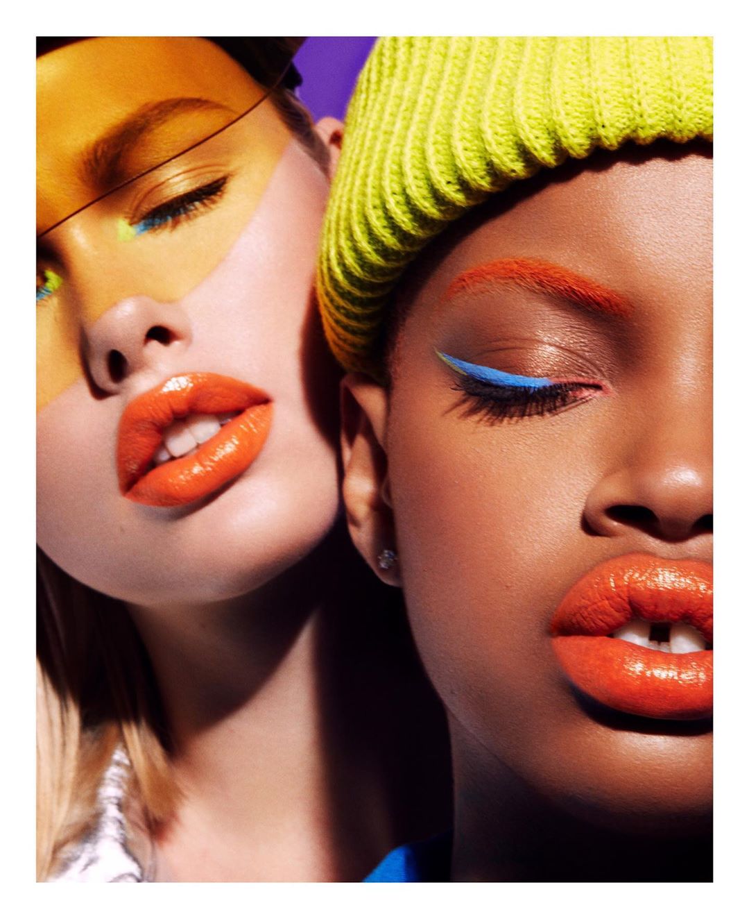 Close-up of two diverse women with vibrant makeup featuring orange lips and colorful eyeliner, one wearing a yellow beanie against a purple backdrop.