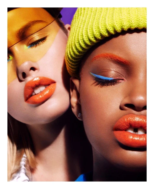 Close-up of two diverse women with vibrant makeup featuring orange lips and colorful eyeliner, one wearing a yellow beanie against a purple backdrop.