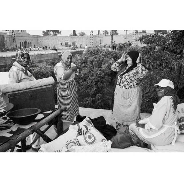 Black and white image of four women in headscarves, aprons, and casual clothing standing outdoors near trees and a building.