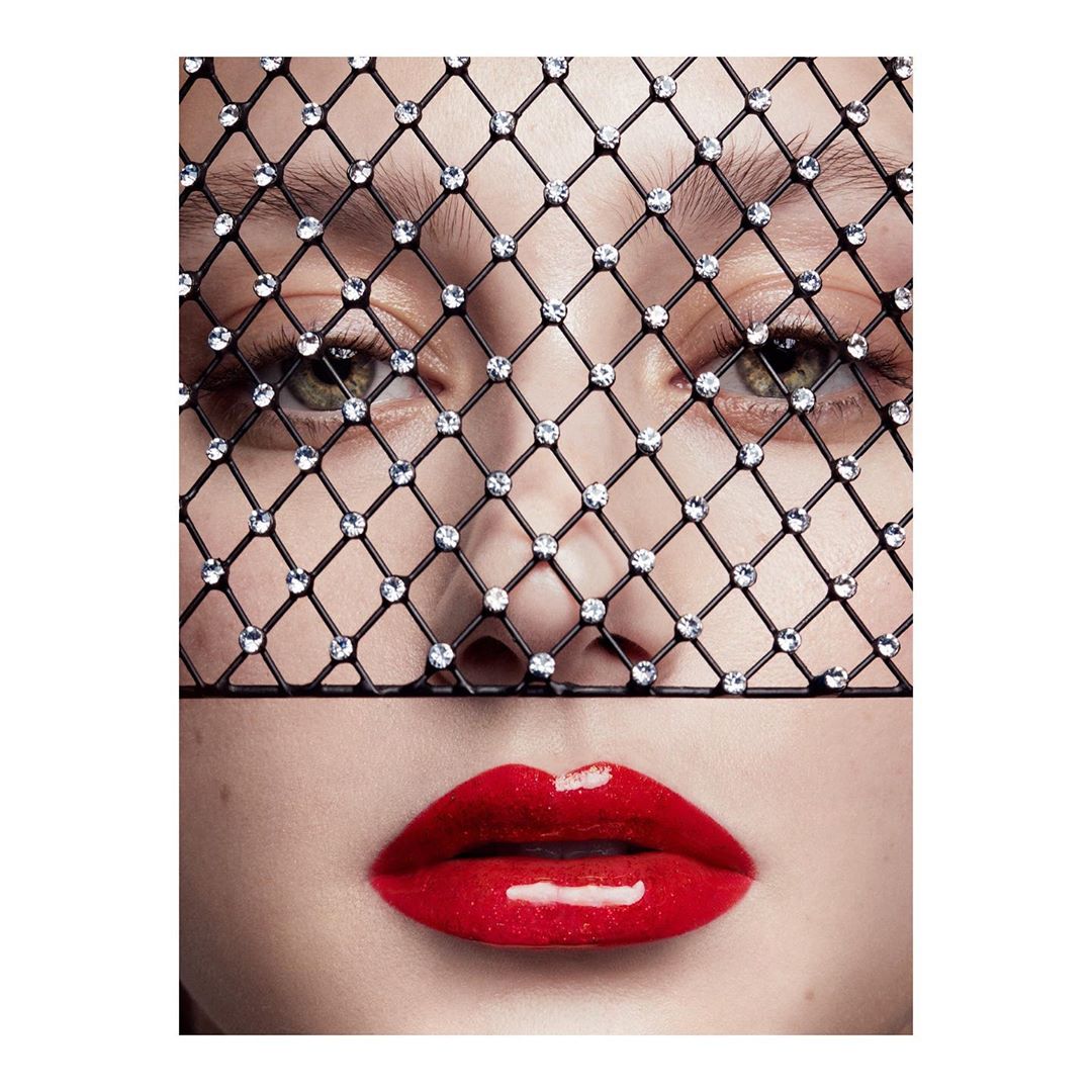 Close-up portrait of a fair-skinned adult female with glossy red lips, wearing a sparkling black mesh face covering, fashion, beauty.