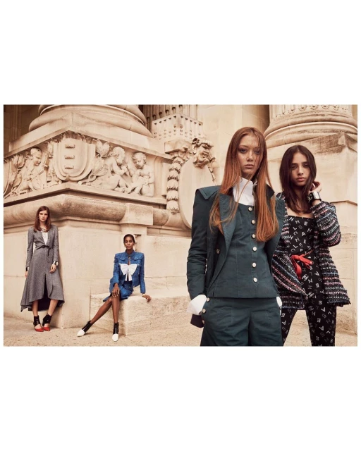 Fashion editorial featuring a group of four female models posing outdoors in front of a beige stone building with carved details.