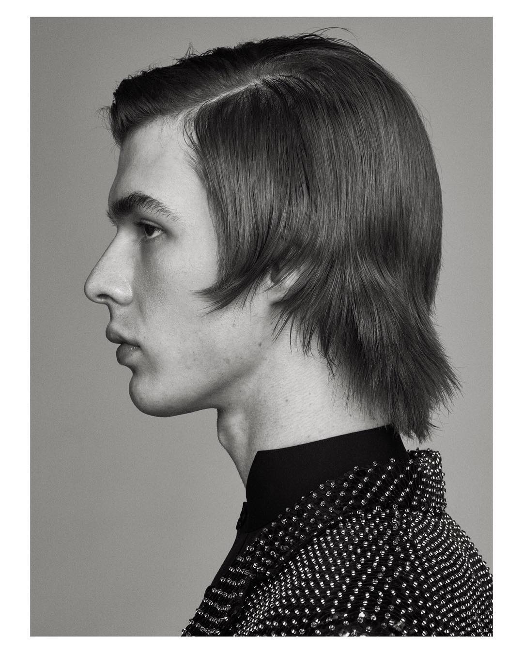 Monochrome portrait of a young man with dark hair in profile, wearing a beaded jacket against a neutral gray background.