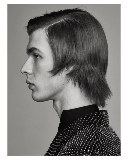 Monochrome portrait of a young man with dark hair in profile, wearing a beaded jacket against a neutral gray background.