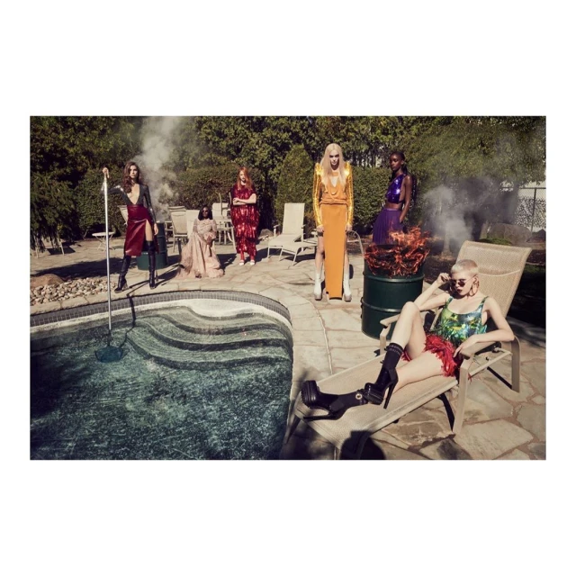 Group of women in colorful dresses pose by a pool surrounded by green trees, a lawn, lounge chairs, and patio furniture outside.