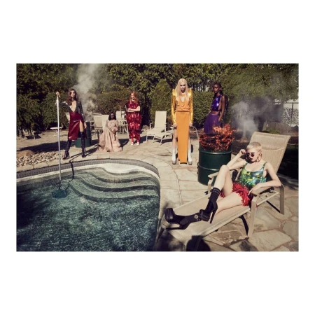 Group of women in colorful dresses pose by a pool surrounded by green trees, a lawn, lounge chairs, and patio furniture outside.