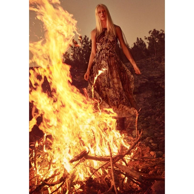 A woman in a patterned dress stands near a bonfire at dusk. The scene combines fashion and natural elements, creating a dramatic image.