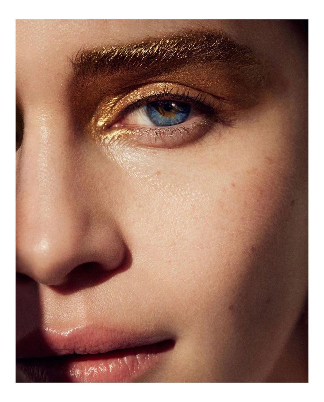 Close-up of a woman's face with golden eyeshadow and blue eyes. Focus on eye makeup and the model's complexion.