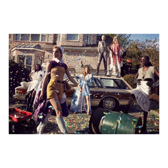 A group of seven fashionably dressed people pose outside a suburban home with a vintage station wagon and confetti falling.