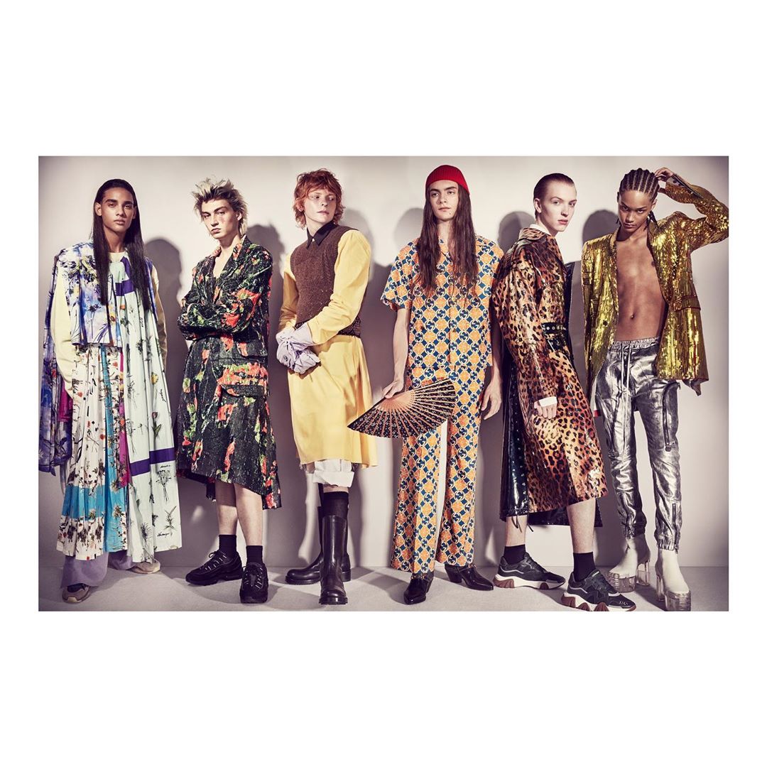 Six fashion models strike a pose in uniquely designed clothing, standing against a neutral backdrop in a fashion studio setting.
