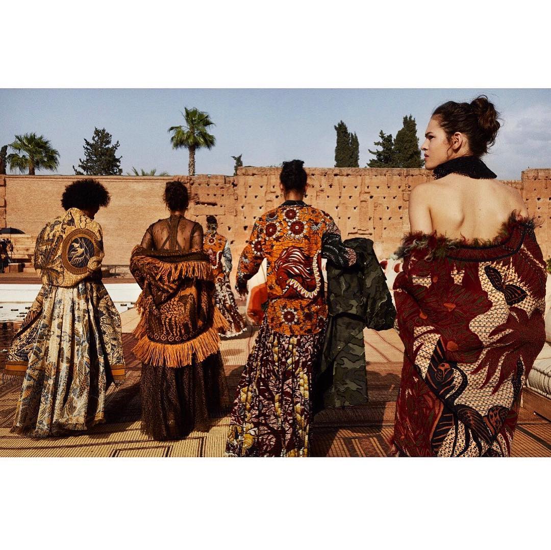 Five fashion models showcasing unique, ornate patterned jackets and skirts outdoors on a sunny day. A building and palm trees appear in the background.
