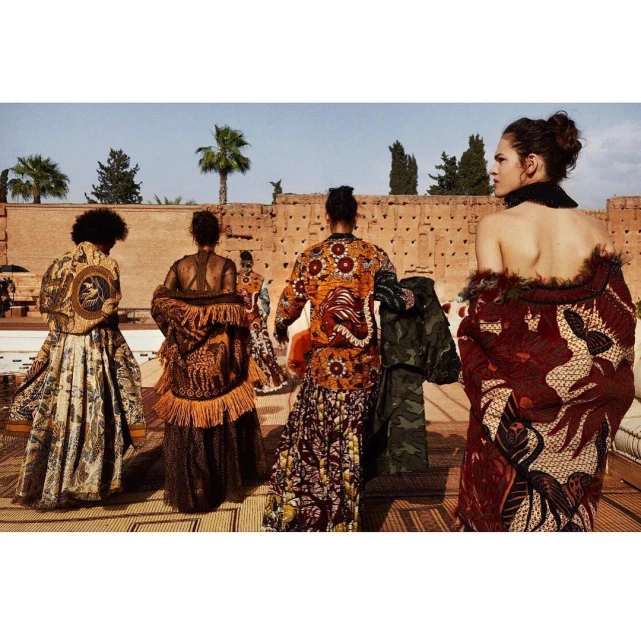 Five fashion models showcasing unique, ornate patterned jackets and skirts outdoors on a sunny day. A building and palm trees appear in the background.