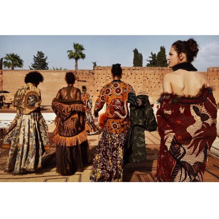 Five fashion models showcasing unique, ornate patterned jackets and skirts outdoors on a sunny day. A building and palm trees appear in the background.