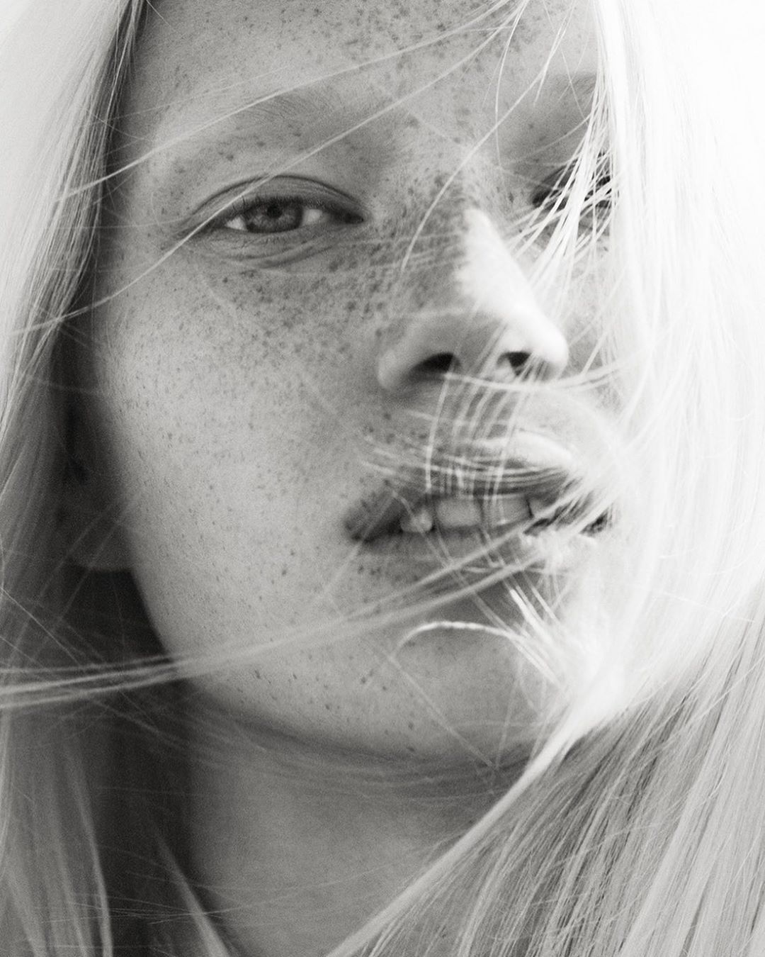 Close-up black and white portrait of a young adult woman with blonde hair and freckles, gazing directly at the camera in a bright, soft light.