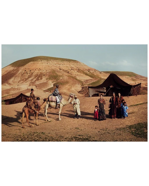 Fashion shoot in desert landscape with models and children gathered near camels and tents against backdrop of sand dunes and clear sky.