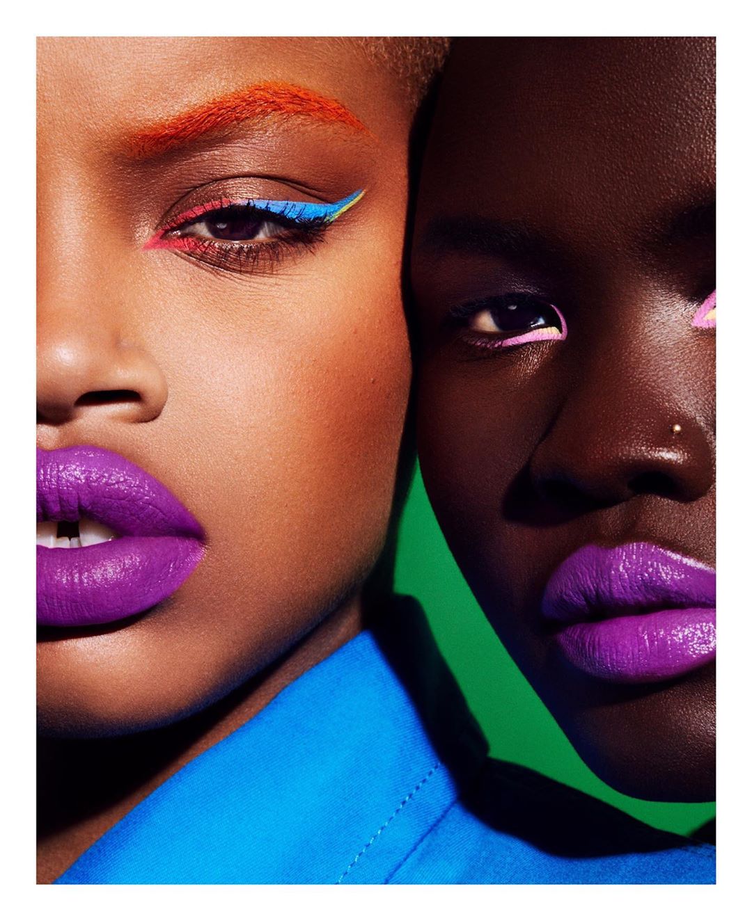Close-up of two women with bold makeup: purple lipstick, orange eyebrows, and colorful eyeliner against a green and blue background.