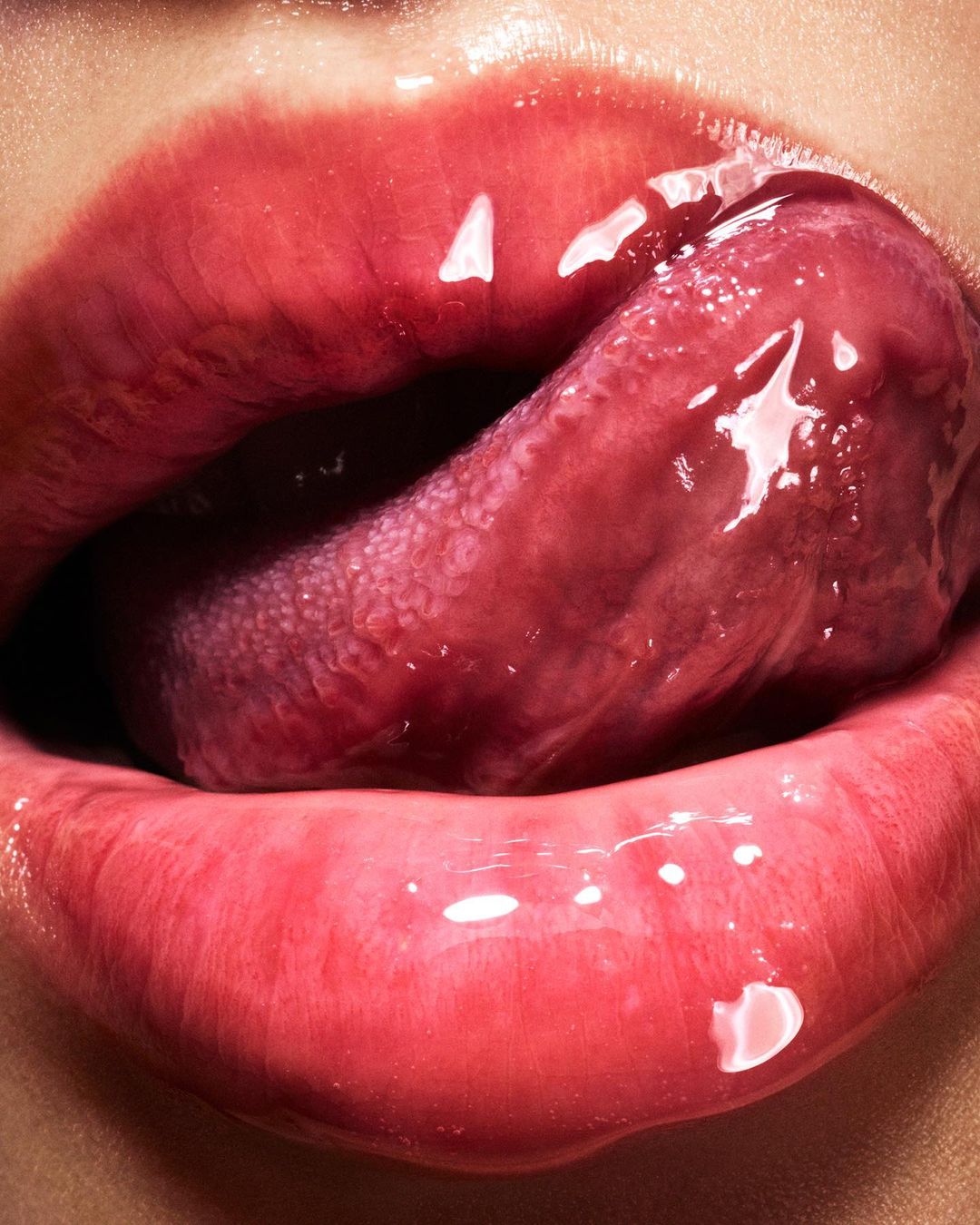 Close-up studio photograph of female lips with glossy lipstick, and a tongue sticking out. The color is red and pink.