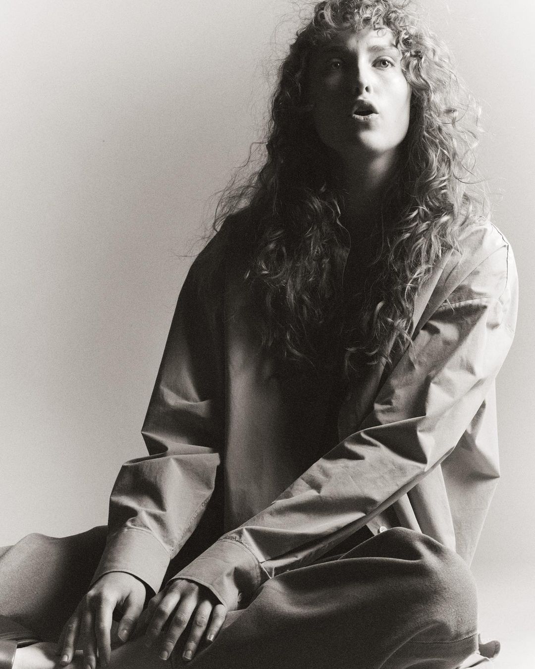 Monochrome portrait of a woman with long curly hair wearing a button-down shirt and trousers seated on a seamless backdrop