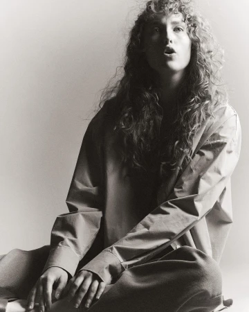 Monochrome portrait of a woman with long curly hair wearing a button-down shirt and trousers seated on a seamless backdrop