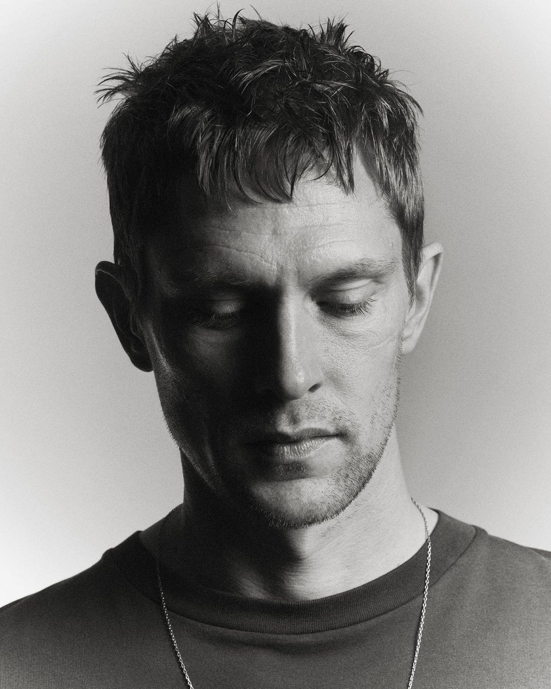 Monochrome portrait of adult male with short hair looking down, wearing a t-shirt and necklace, with soft, diffused lighting.