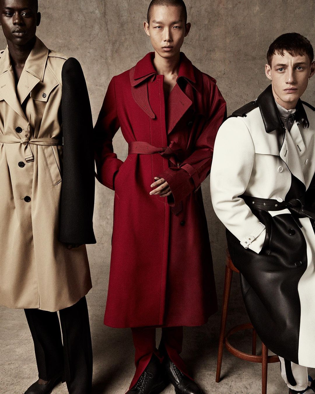 Three fashion models in trench coats stand in front of a neutral backdrop. A man in a red coat is centered, flanked by two others.