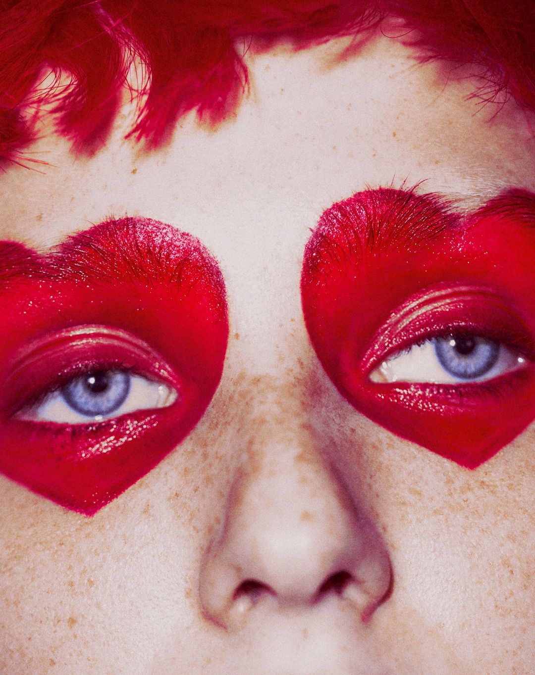 Extreme close-up on an adult female face with red heart-shaped makeup around the eyes and freckles on her nose. Red hair is visible.