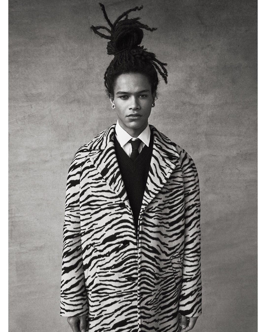 Portrait of a young adult male with dreadlocks, wearing a zebra-striped coat, a sweater, and a tie, standing against a neutral background.