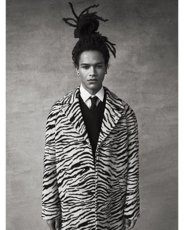 Portrait of a young adult male with dreadlocks, wearing a zebra-striped coat, a sweater, and a tie, standing against a neutral background.