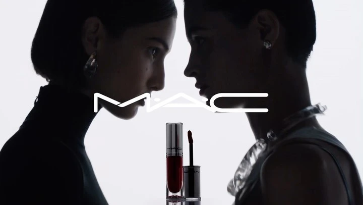 Two adult females and a lipstick product in a beauty-focused shot with a white background and the MAC Cosmetics logo.