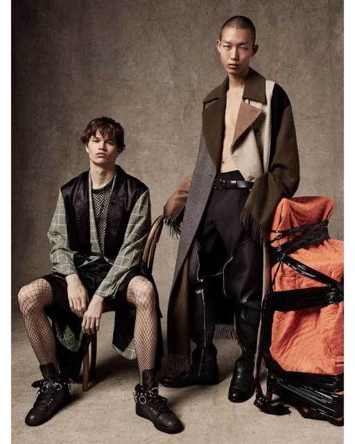 Two young adult fashion models pose indoors in avant-garde outfits; one sits in a chair, the other stands by an orange wrapped object.