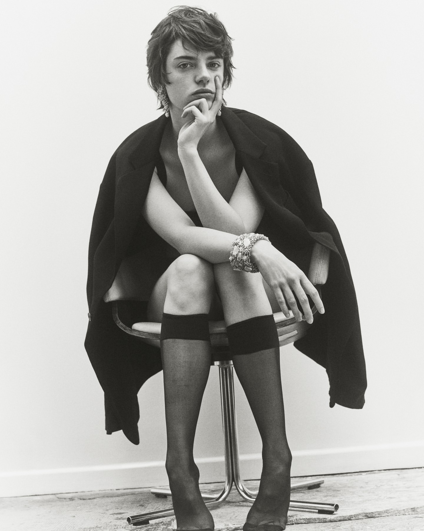 Monochrome portrait of androgynous individual in dark jacket, seated on chrome stool in black stockings, wearing ornate jewelry.