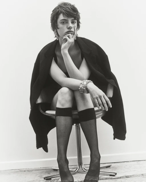 Monochrome portrait of androgynous individual in dark jacket, seated on chrome stool in black stockings, wearing ornate jewelry.