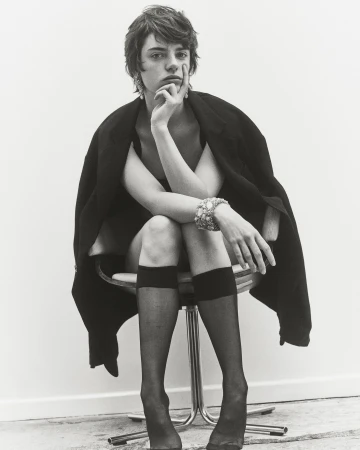 Monochrome portrait of androgynous individual in dark jacket, seated on chrome stool in black stockings, wearing ornate jewelry.