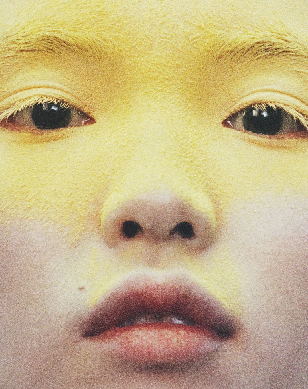 Close-up portrait of a person's face covered in yellow powder, highlighting their eyes, nose, and lips. The composition is simple and minimalist.