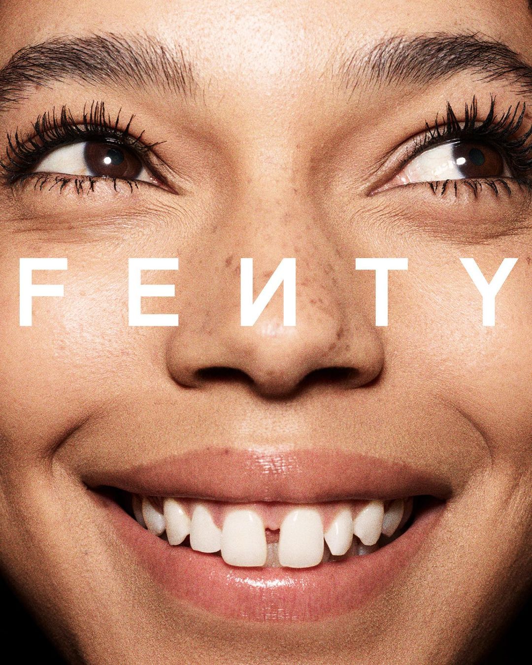 Close-up beauty shot of an adult female face featuring a gap-toothed smile, dark eyes, and fair skin, set against a dark background.