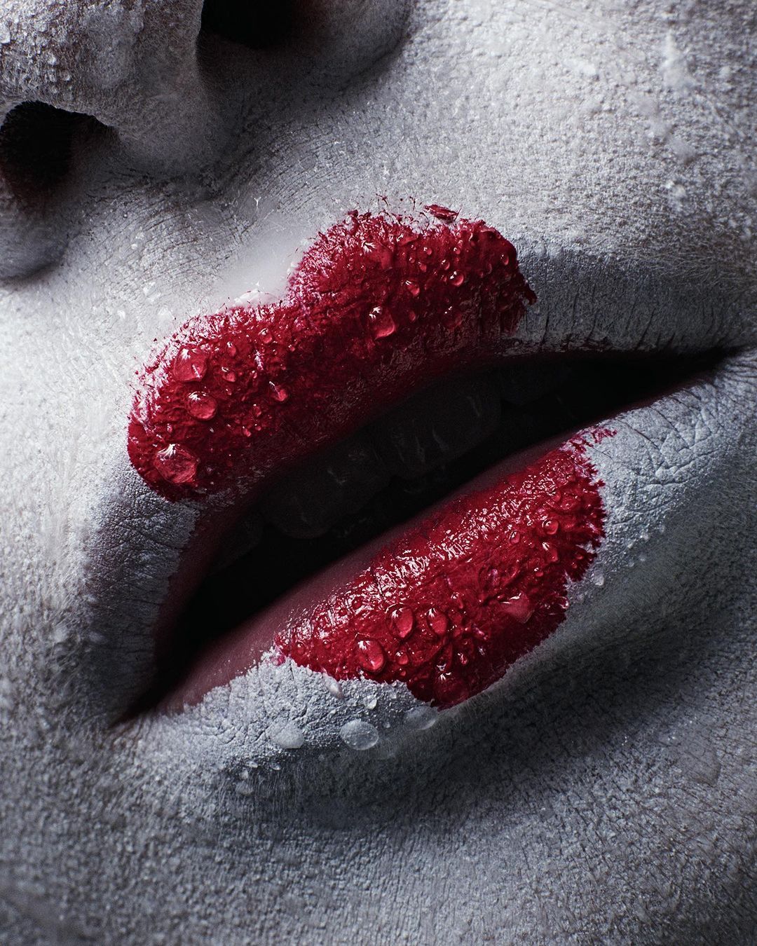 Extreme close-up shot of red lipstick on a woman's mouth, her face covered in greyish-white powder, with water droplets on her skin.