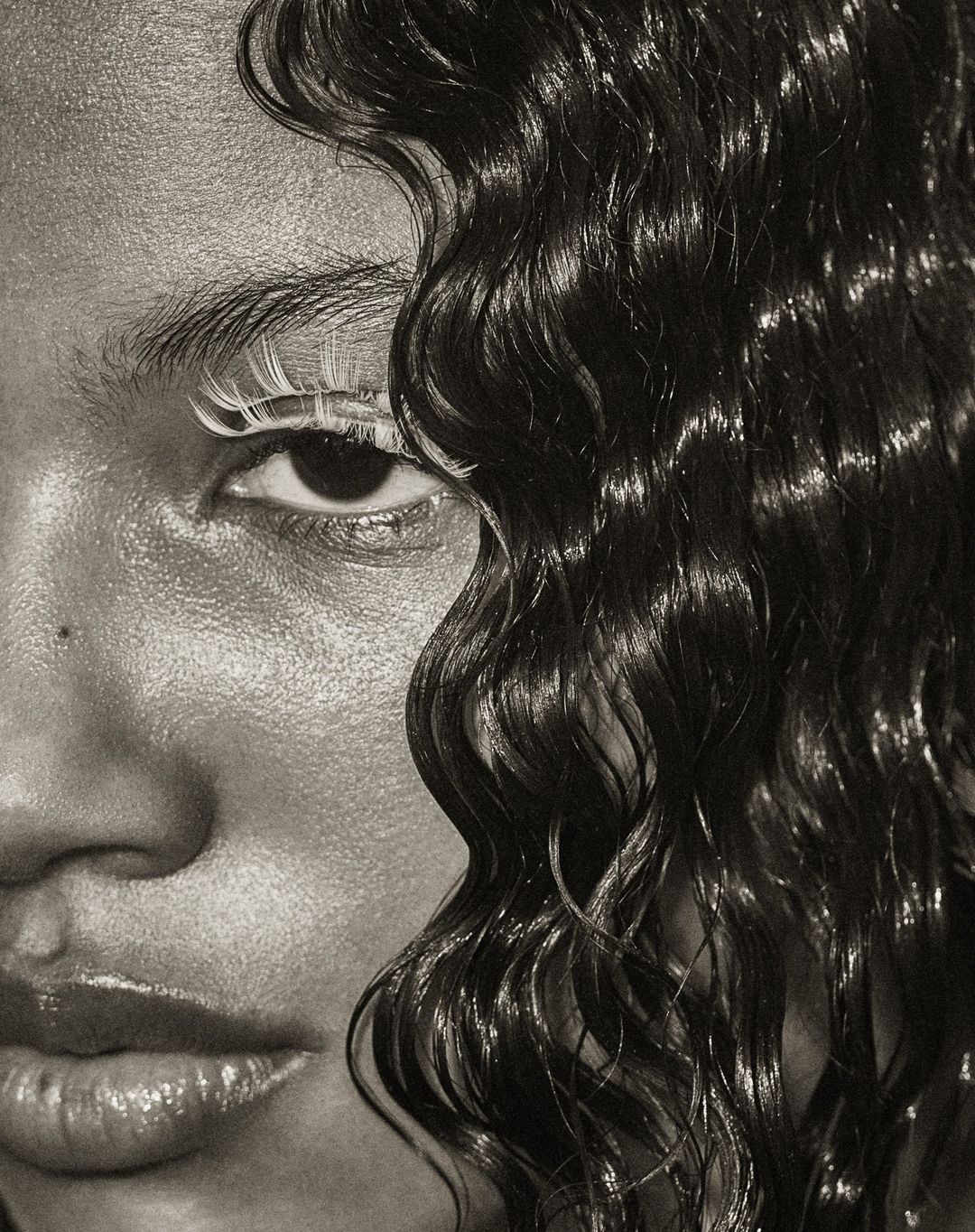 Close-up portrait of a Black female model with glossy skin, wet wavy hair, and striking white eyelashes, against a neutral backdrop.