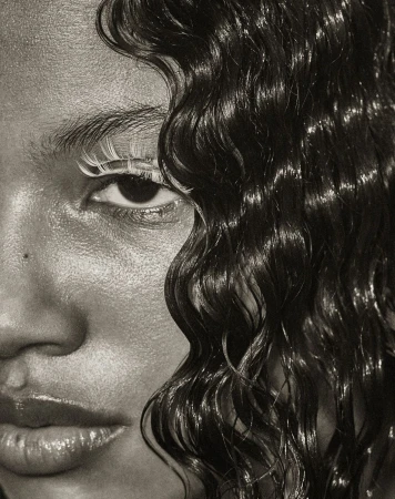 Close-up portrait of a Black female model with glossy skin, wet wavy hair, and striking white eyelashes, against a neutral backdrop.