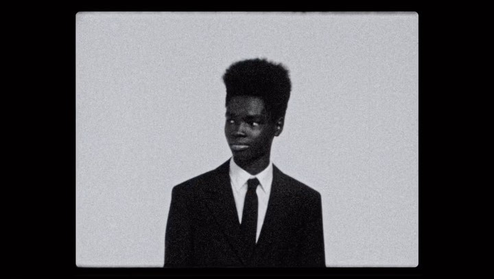 Portrait of a black man wearing a dark suit and tie against a plain light background, captured with heavy film grain.