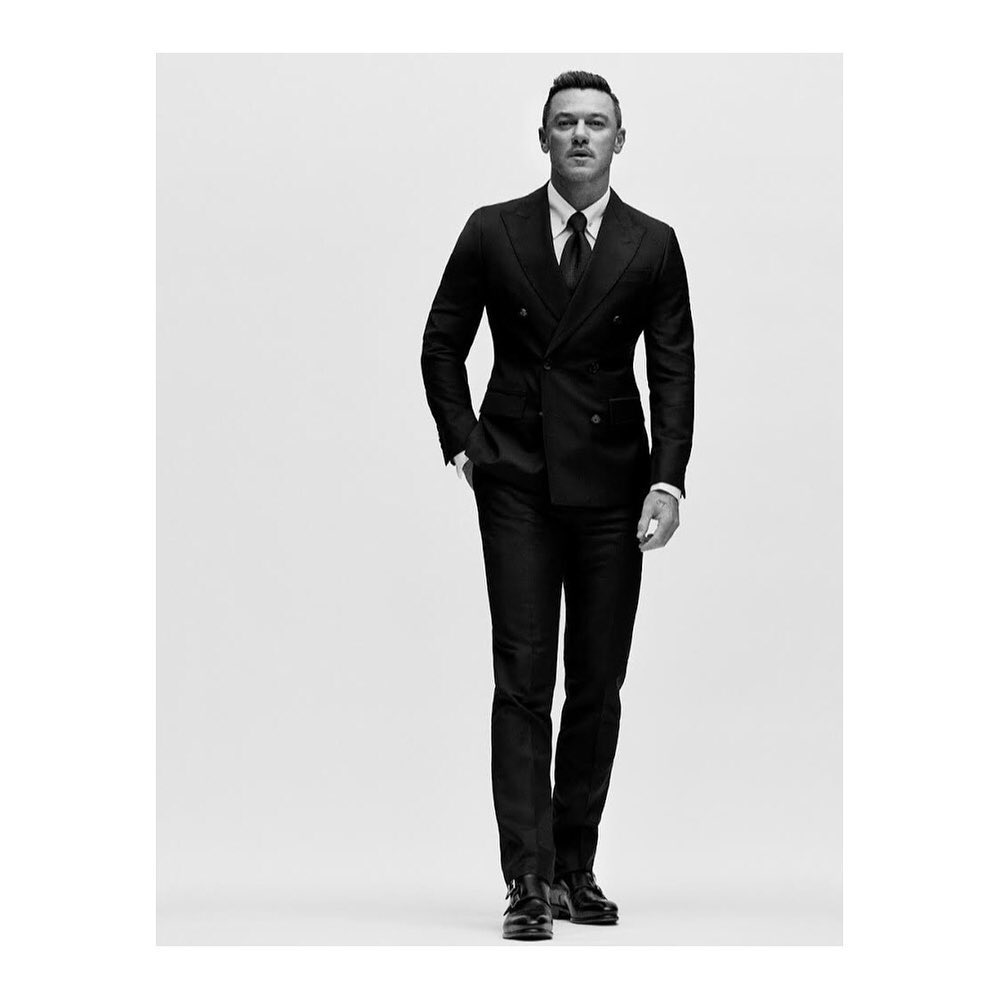 Man wearing a black double-breasted suit and dark tie against a white seamless studio backdrop.