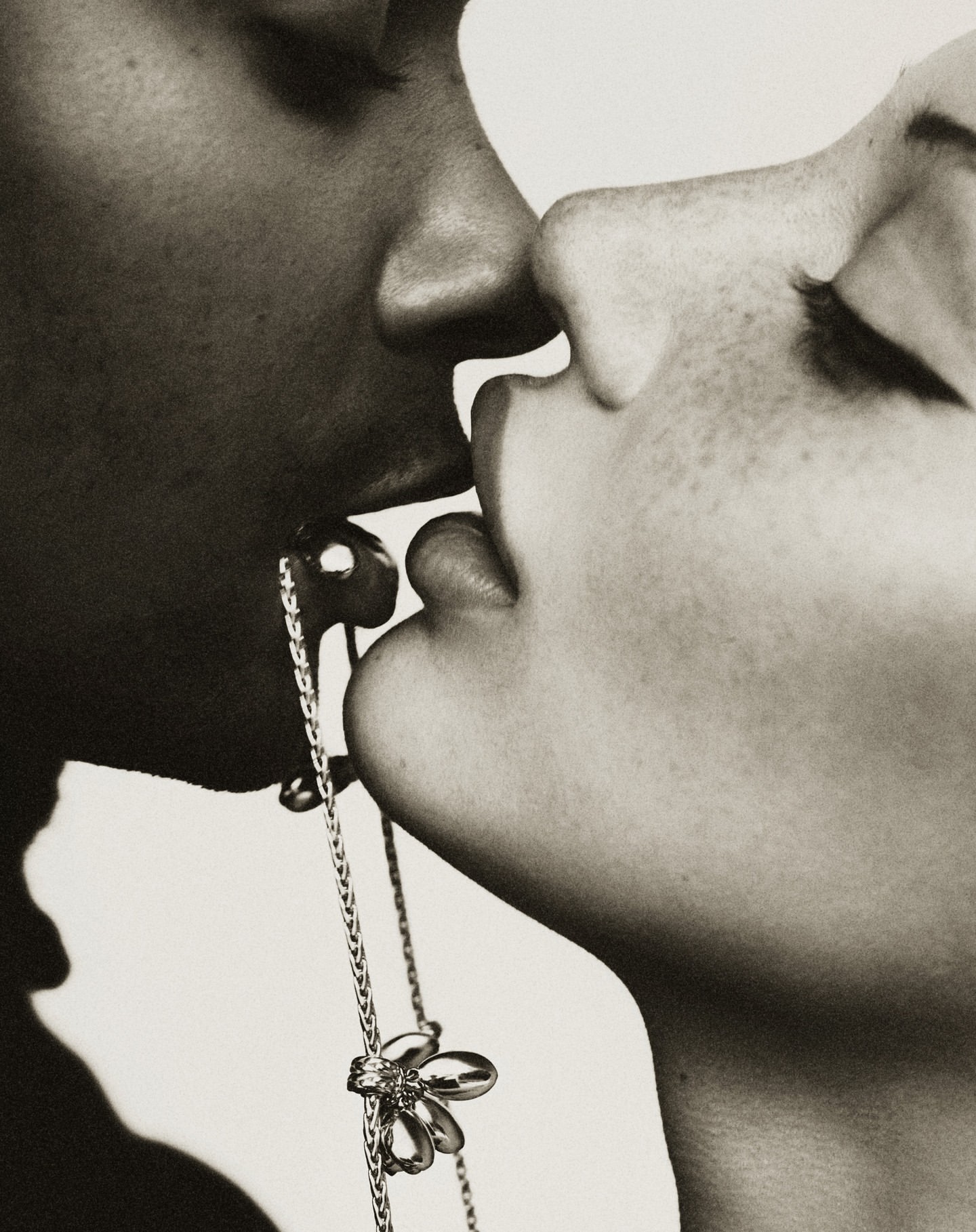 Close-up of two adults kissing, featuring a gold chain earring with a flower detail. Black and white photograph, faces in frame.